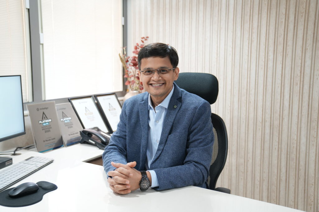 Dr. Ronak Joshi, MDS Orthodontist, sitting smiling at his consultation desk with his Platinum Elite Invisalign Provider awards proudly on display at Anant Dental Clinic in Gurukul, Ahmedabad.